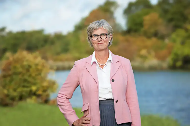 Bettina Lundgren i hvid skjorte og lyserød blazer, med hånd på hoften og smil uden tænder. Billedet er beskåret ved hoften. Baggrund er natur med træer og sø.